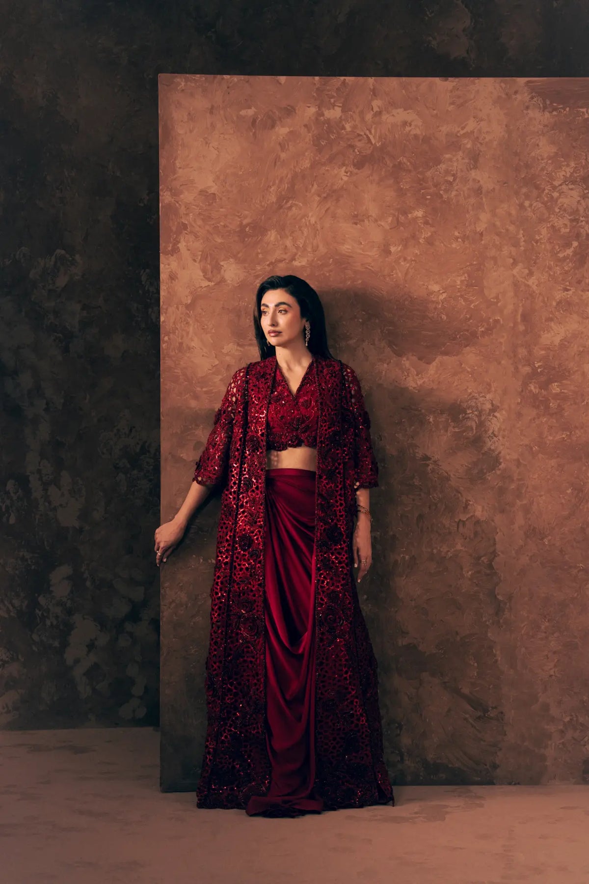 Model leaning on backdrop in Crimson Pakistani formal wear with open sequin jacket, blouse and satin silk skirt