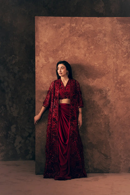Model leaning on backdrop in Crimson Pakistani formal wear with open sequin jacket, blouse and satin silk skirt